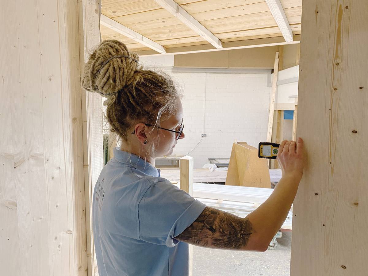Eine Person misst mit einem Lasermessgerät in einem Holzbau. Sie trägt ein blaues Hemd und hat einen Dutt. Im Hintergrund sind Holzmaterialien.