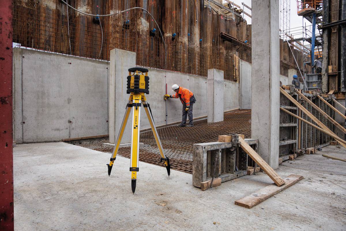 Ein Bauarbeiter in orangefarbener Schutzkleidung vermisst eine Baustelle mit einem Vermessungsgerät im Vordergrund.