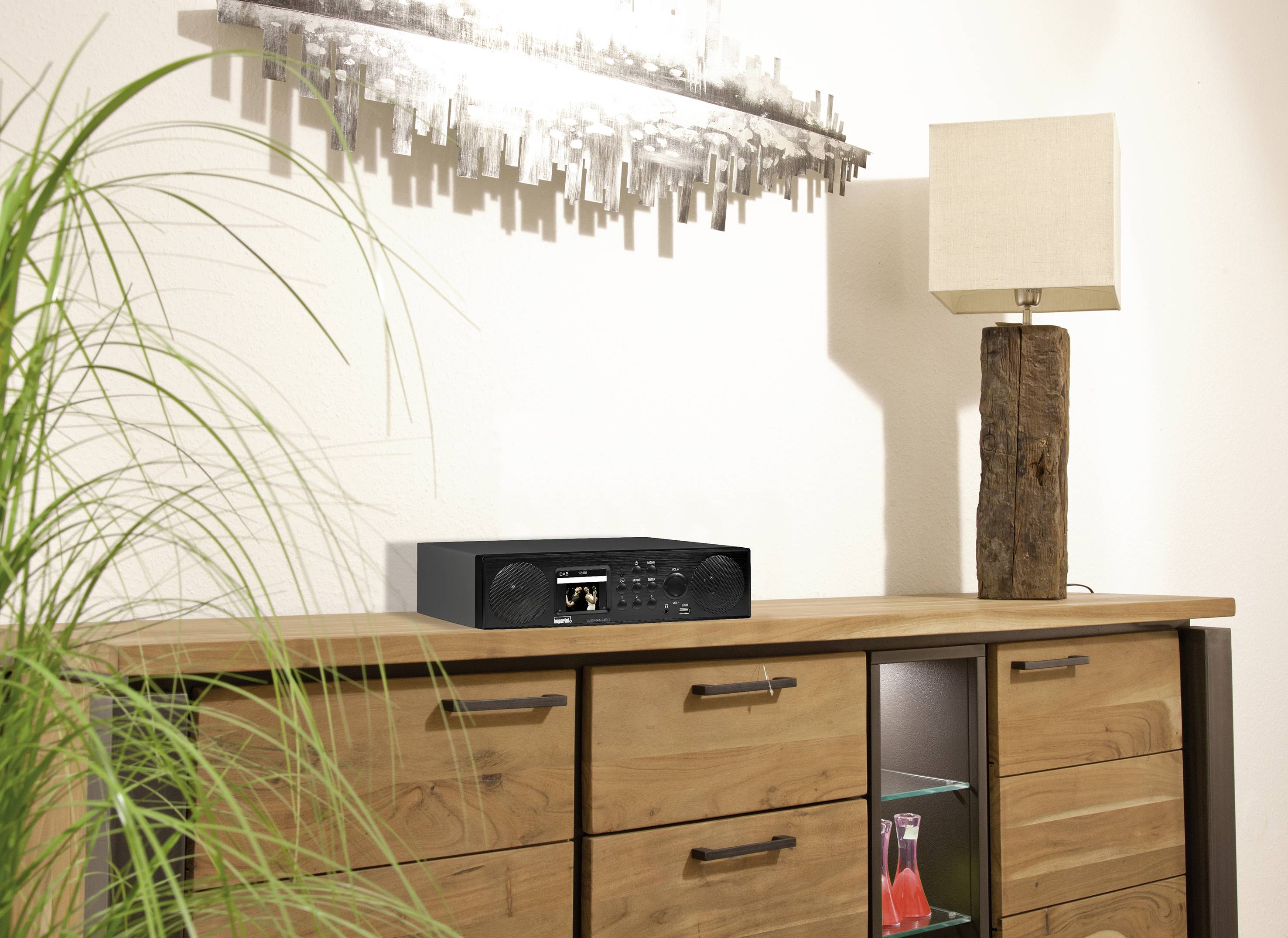 'Tisch mit Holzschubladen, darauf eine Musikanlage und eine Lampe. An der Wand hängt ein dekoratives Metallschild. Eine Pflanze im Vordergrund.'