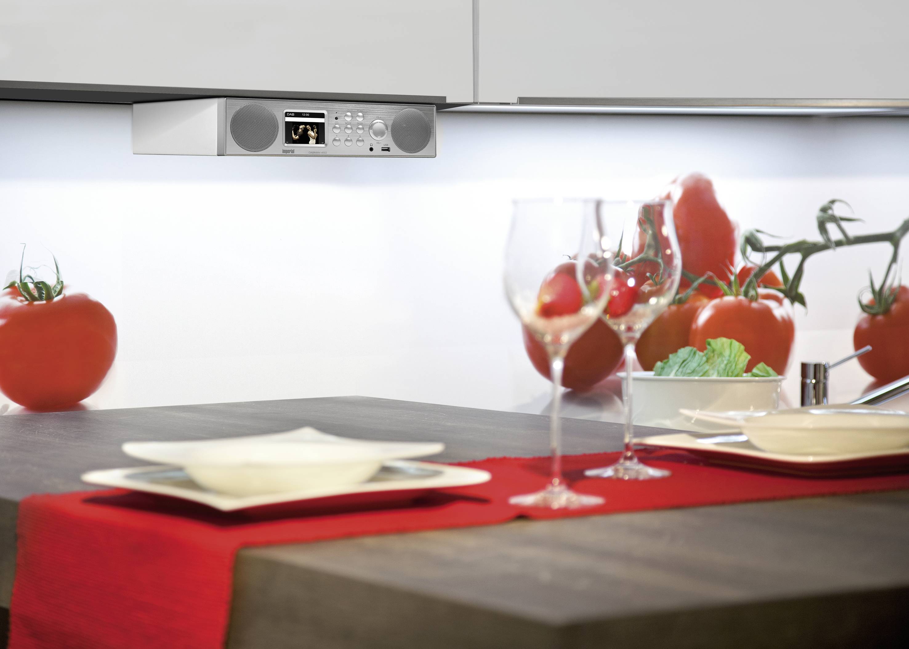 Küchentisch mit rotem Läufer, gedeckt für zwei. Tomaten im Hintergrund; an der Wand ein Radio mit Display und Lautsprechern.