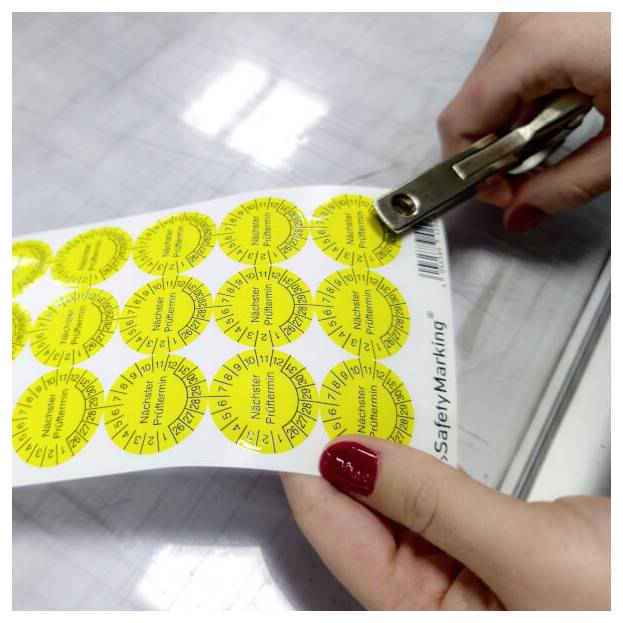 Eine Person verwendet einen Locher an einem Blatt gelber, runder Etiketten mit Zahlen und Monaten, beschriftet mit „SafetyMarking".