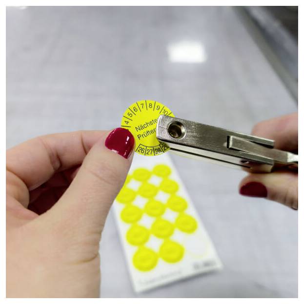 Eine Hand benutzt einen Locher, um einen gelben Aufkleber mit Zahlen auf einem Blatt zu markieren. Der Fokus liegt auf der Zahl '7', was eine Auswahl oder Aktion andeutet.