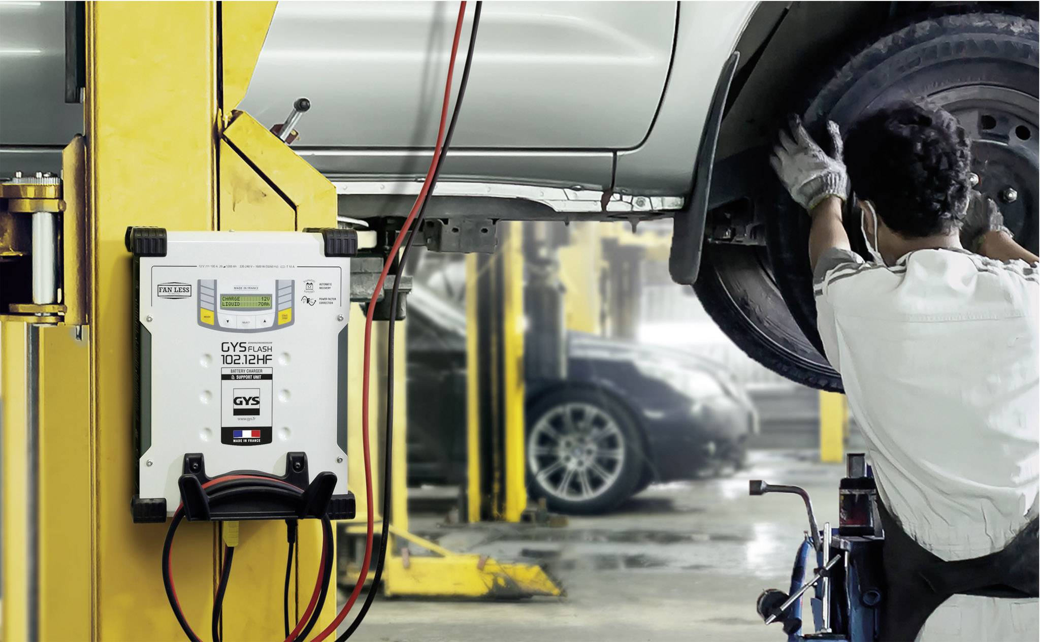 Ein Mechaniker inspiziert ein aufgebocktes Auto in einer Werkstattumgebung. Im Vordergrund ist ein Batterieladegerät zu sehen.