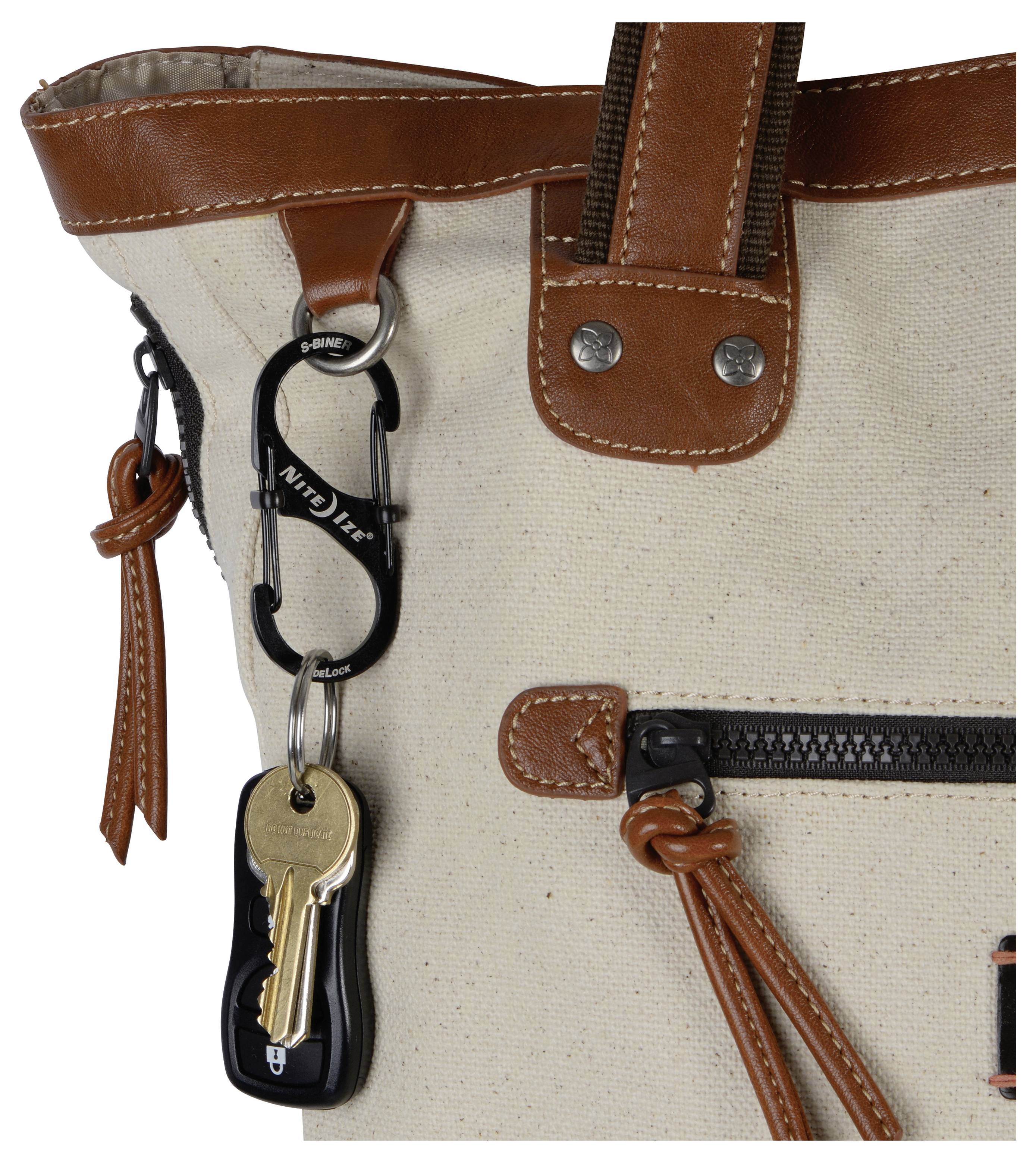 Eine beige Tasche mit braunen Lederriemen und Reißverschlüssen hat einen schwarzen Karabiner, an dem zwei Schlüssel und ein schwarzer Autoschlüsselanhänger befestigt sind.