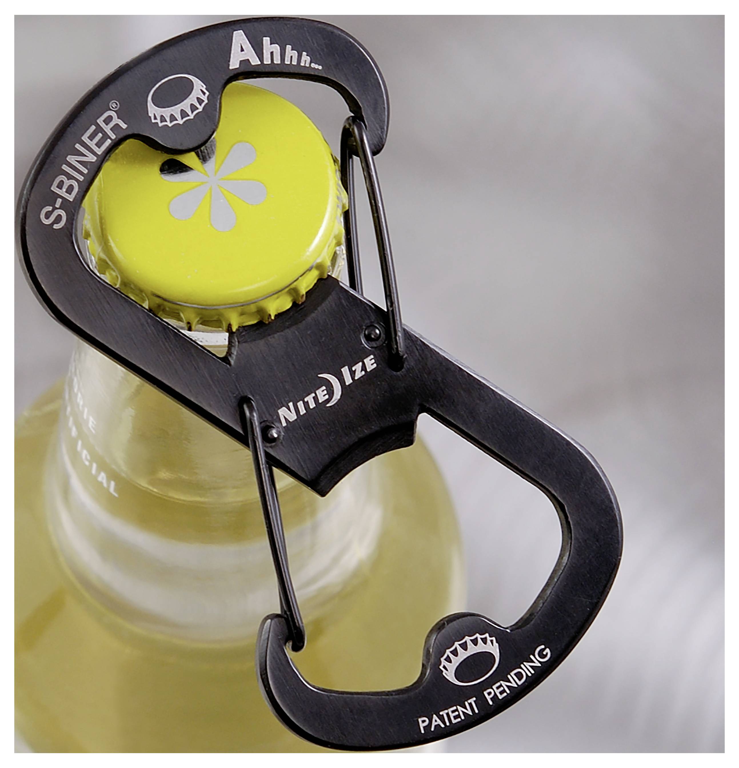 Das Bild zeigt einen schwarzen S-Biner-Flaschenöffner mit der Aufschrift "Ahhh..." und "Patent angemeldet", der an einem gelben Kronkorken einer Glasflasche befestigt ist.