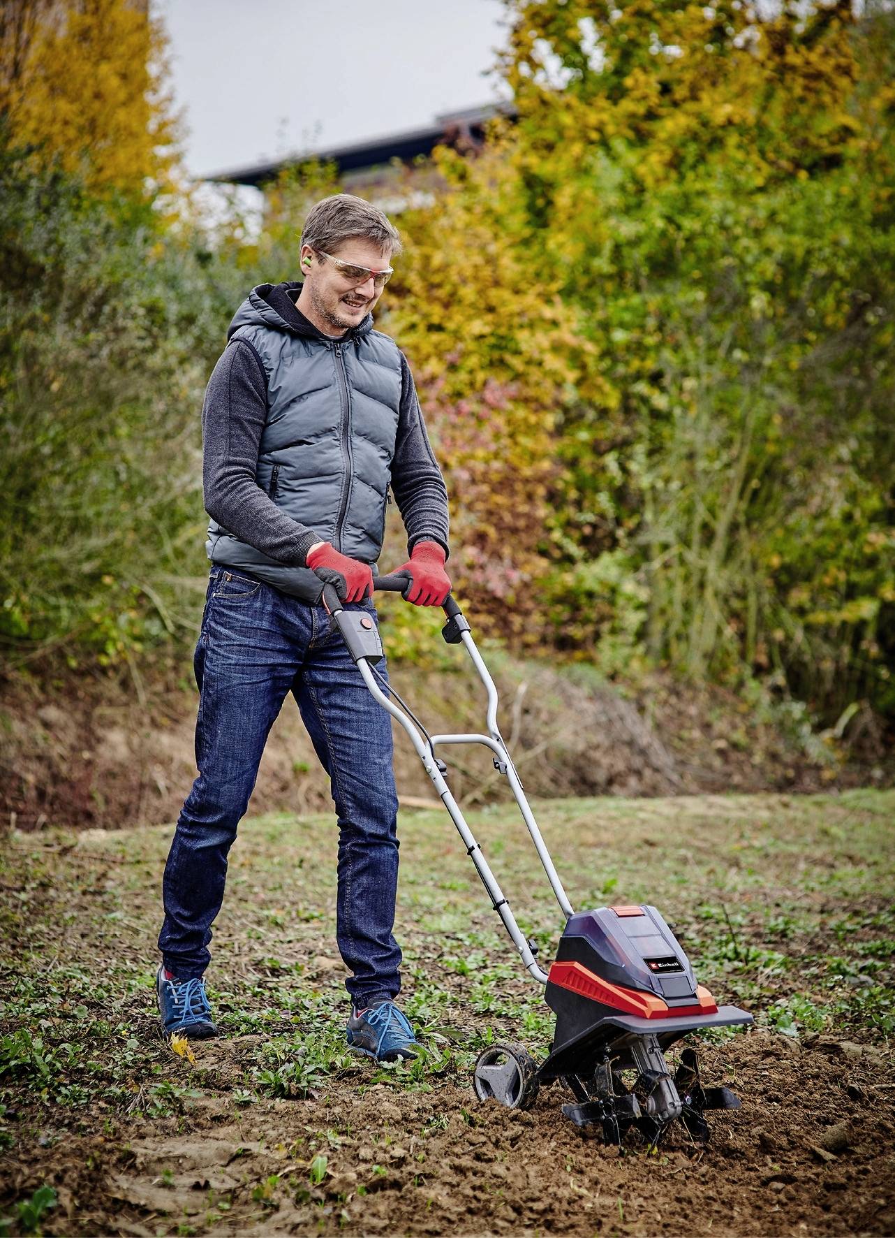 Eine Person in Freizeitkleidung bedient eine rote Gartenfräse auf einem Feld mit herbstlicher Vegetation im Hintergrund.