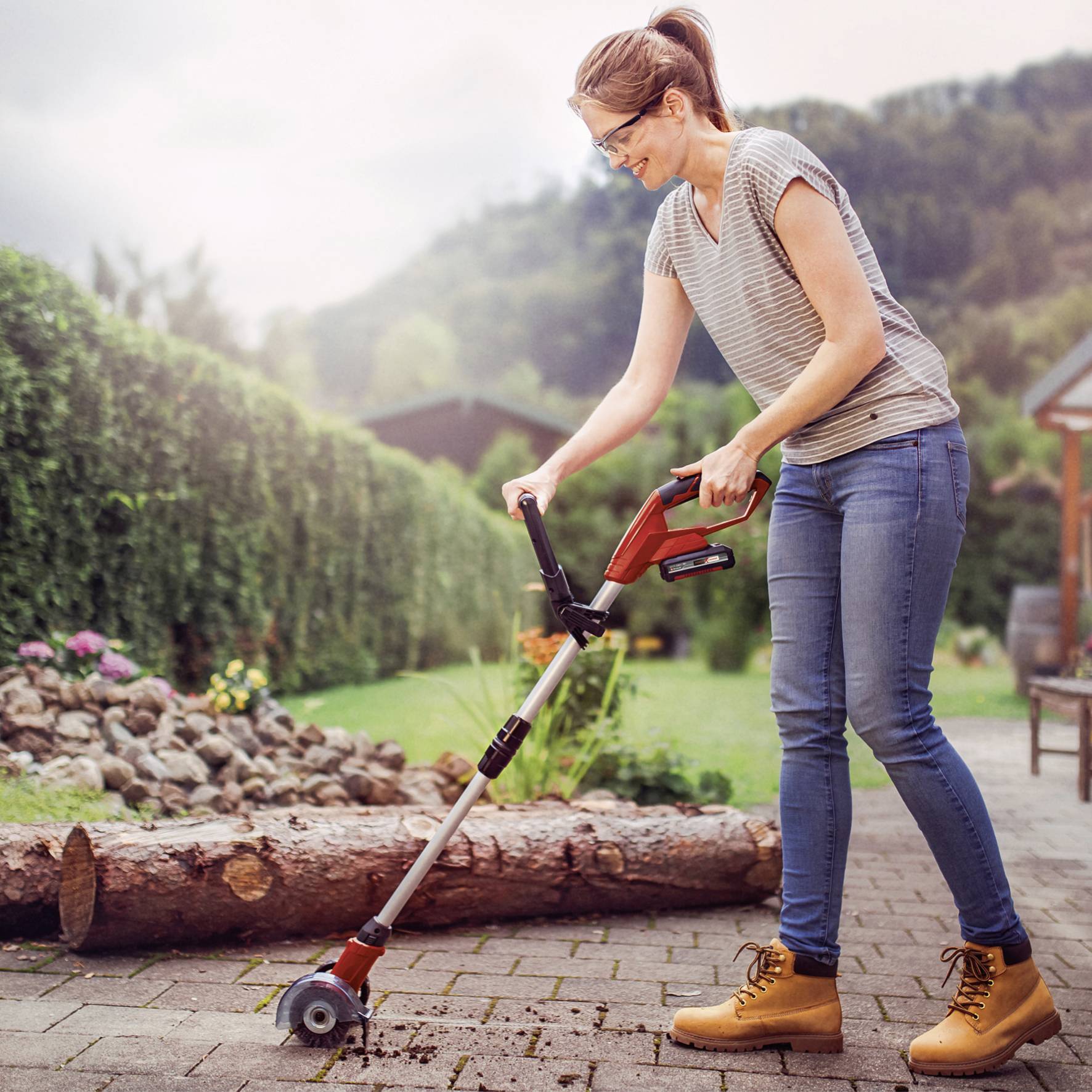Eine Frau mäht den Rasen mit einem elektrischen Trimmer auf einer Terrasse. Im Hintergrund Bäume und Berge an einem sonnigen Tag.