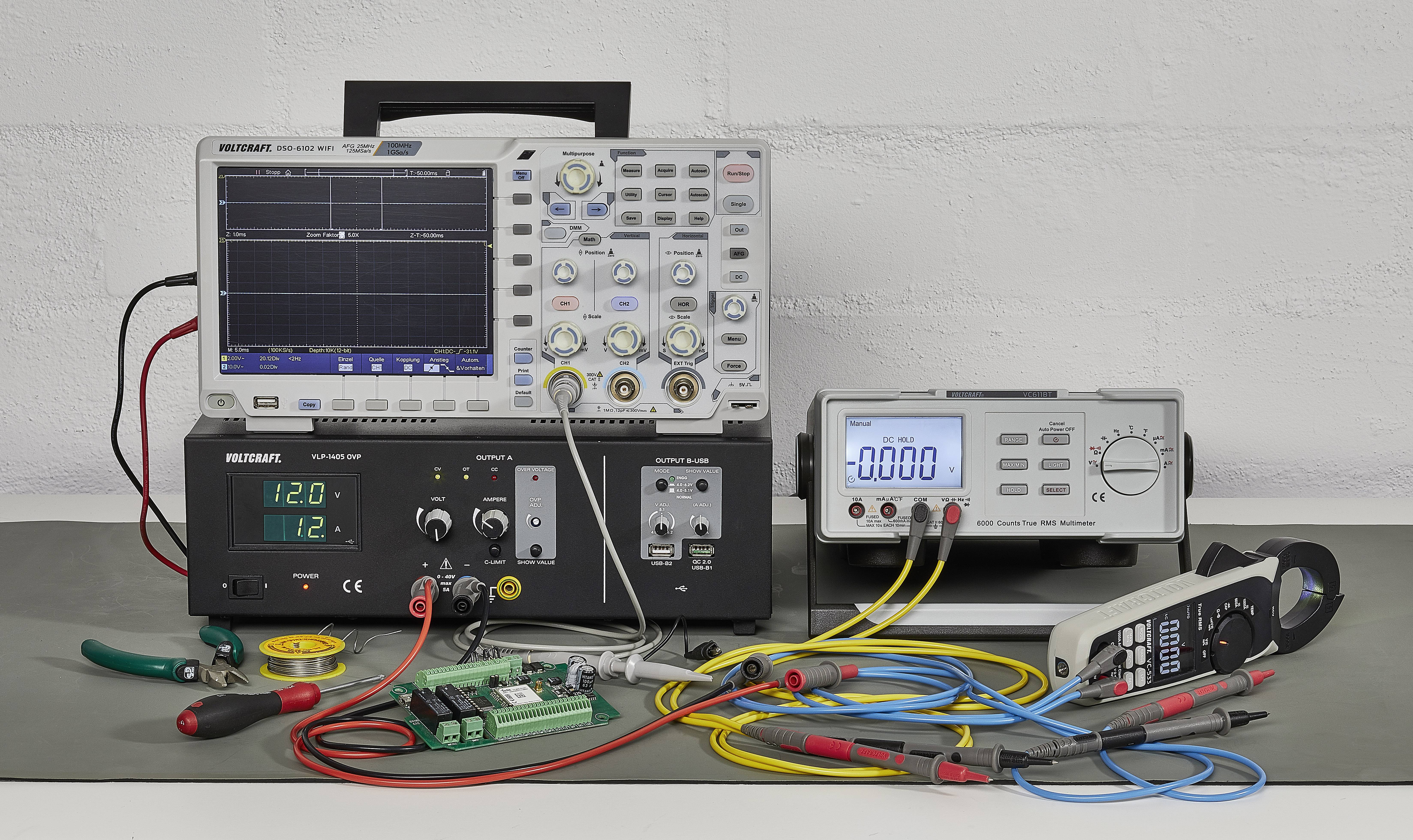 Ein Tisch mit elektronischen Messgeräten: Oszilloskop, Stromversorgungsgeräte, Digitalmultimeter. Kabel und Werkzeug liegen bereit.