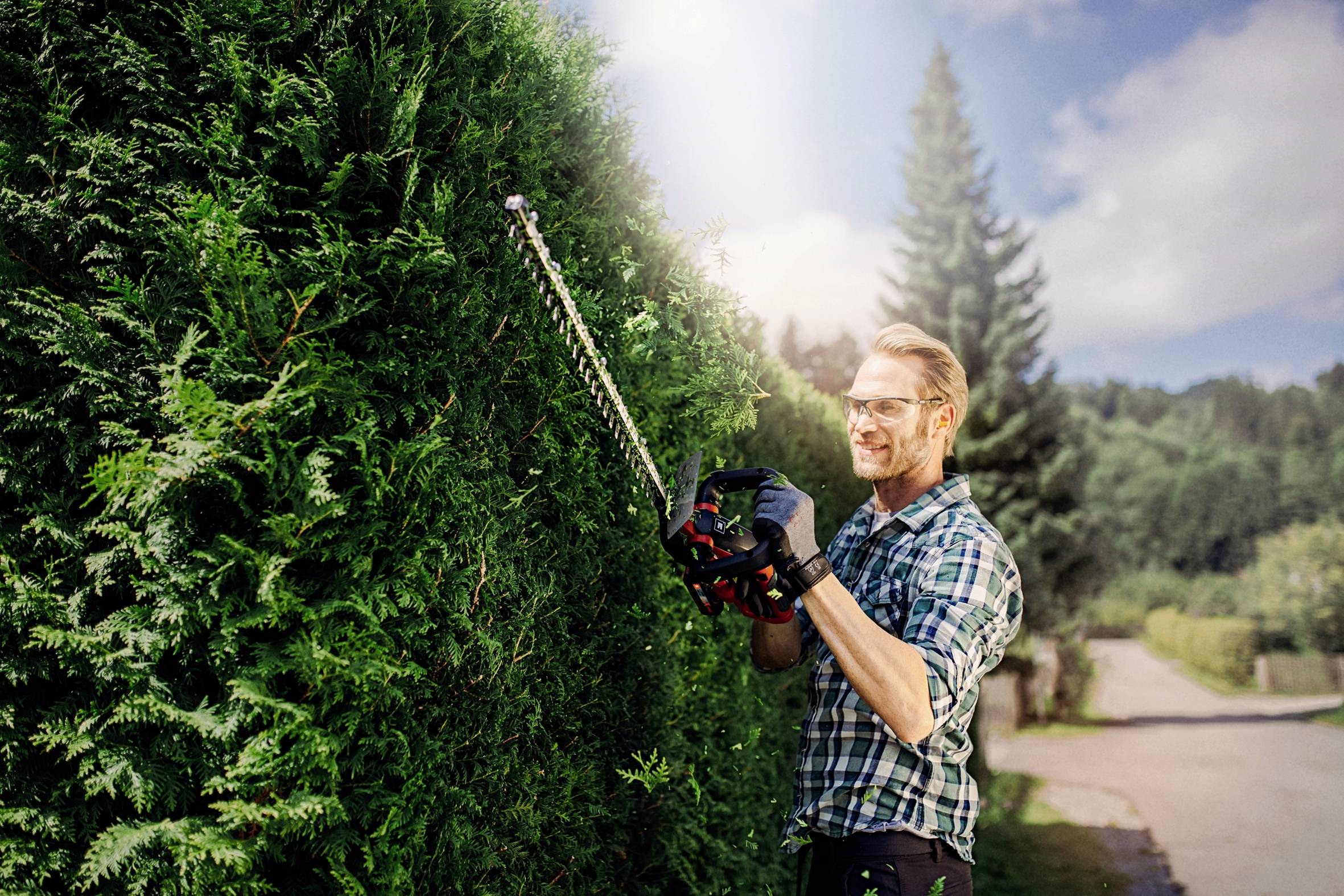 Ein Mann in kariertem Hemd schneidet mit einer Heckenschere eine hohe Hecke. Im Hintergrund ein sonniger Tag mit Bäumen.