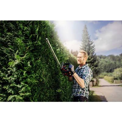 Ein Mann in kariertem Hemd schneidet mit einer Heckenschere eine hohe Hecke. Im Hintergrund ein sonniger Tag mit Bäumen.