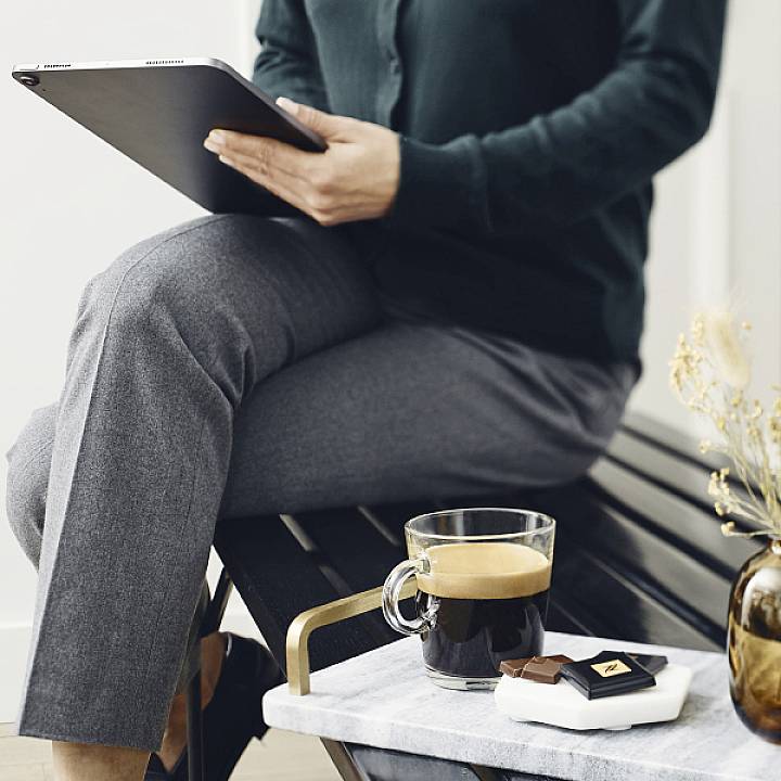 Eine sitzende Person hält ein Tablet in den Händen. Auf einem Tisch vor ihr stehen eine Tasse Kaffee und ein Teller mit Schokolade.