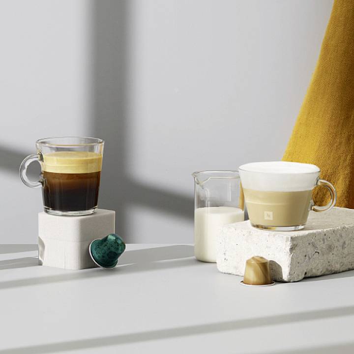 Zwei Tassen Kaffee auf Steinen, begleitet von zwei Kaffeekapseln, eine grün und eine gold, in einer minimalistischen Umgebung.