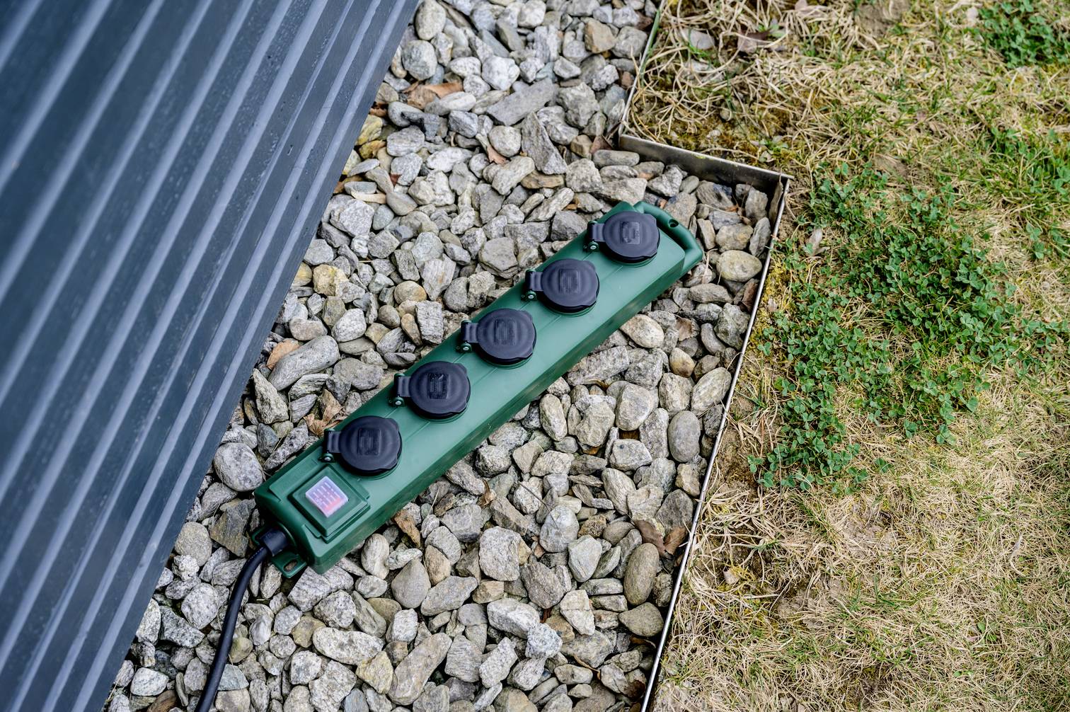 Eine grüne Mehrfachsteckdose mit fünf schwarzen Steckplätzen liegt auf Kiesboden neben einer metallenen Wand und grasbewachsenem Rand.
