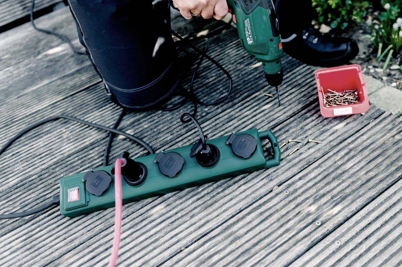 Eine Person auf einem Holzboden benutzt eine grüne Bohrmaschine neben einer Steckdosenleiste und einer roten Box mit Schrauben.