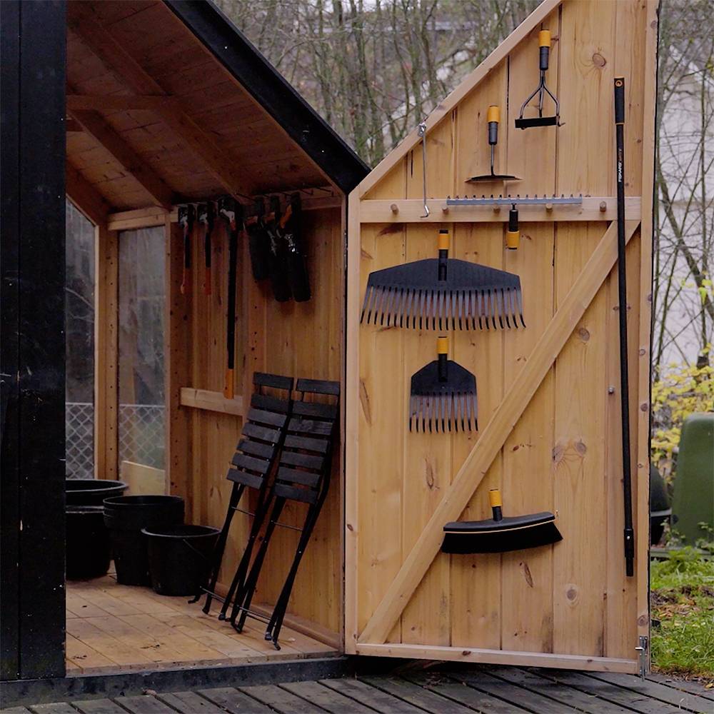 Ein Holzschuppen mit offenem Dach, in dem Gartengeräte wie Rechen, Schaufeln und Stühle ordentlich an den Wänden hängen.