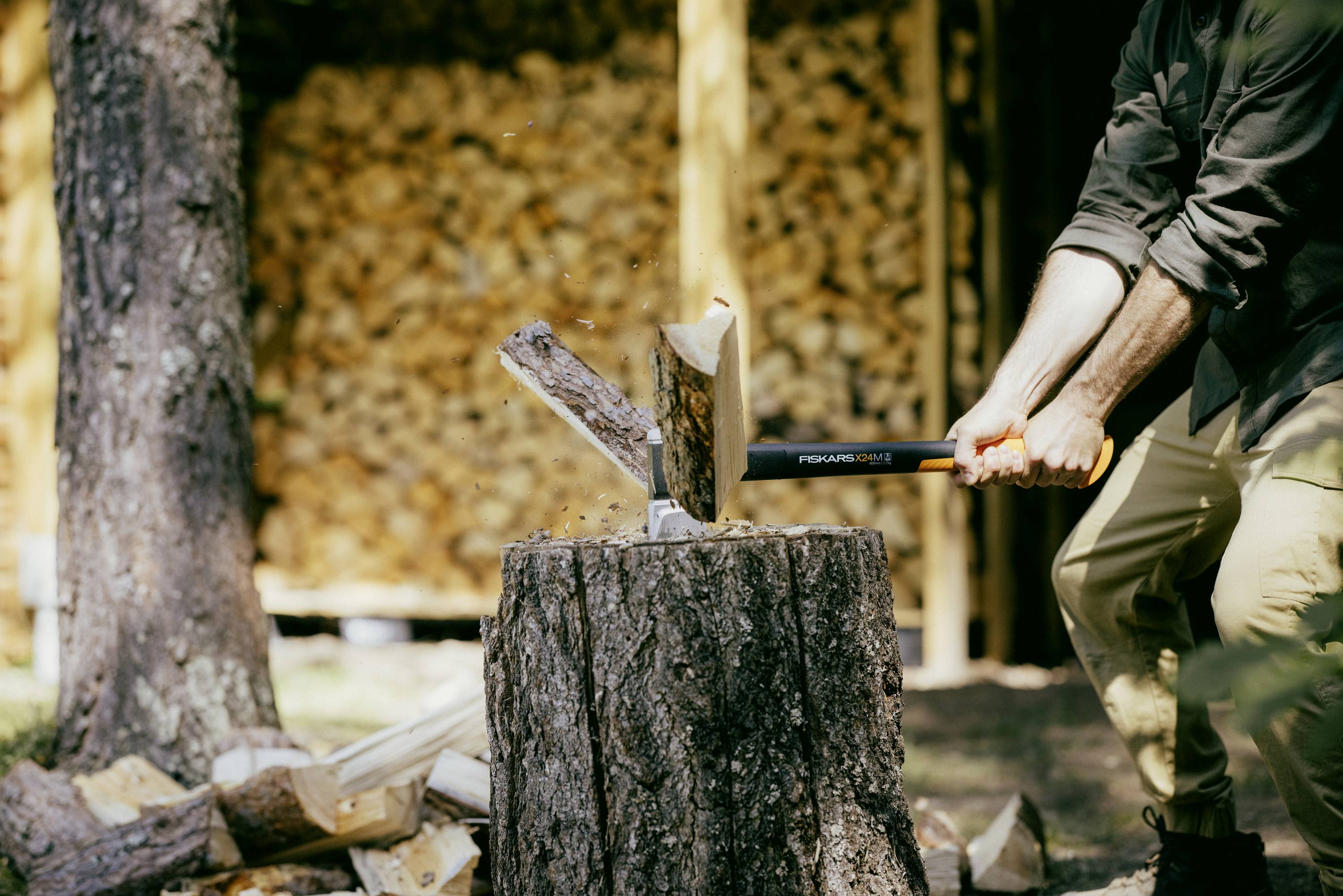 Person spaltet Holz mit einer Axt auf einem Baumstumpf. Im Hintergrund ist ein Stapel geschnittener Holzscheite sichtbar.