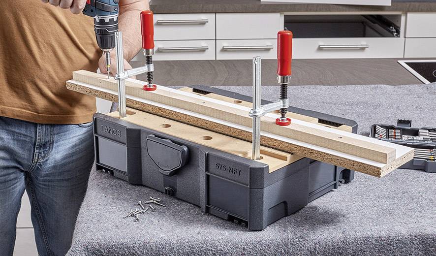 Eine Person befestigt Holzstücke auf einer Werkbank mit Schraubzwingen und nutzt eine Bohrmaschine, um Löcher in das Holz zu bohren.