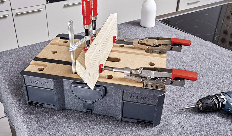 Eine Werkbank mit Holzplatte, die mit Zwingen fixiert ist. Eine Bohrmaschine liegt daneben. Die Szene zeigt einen Werkvorgang.