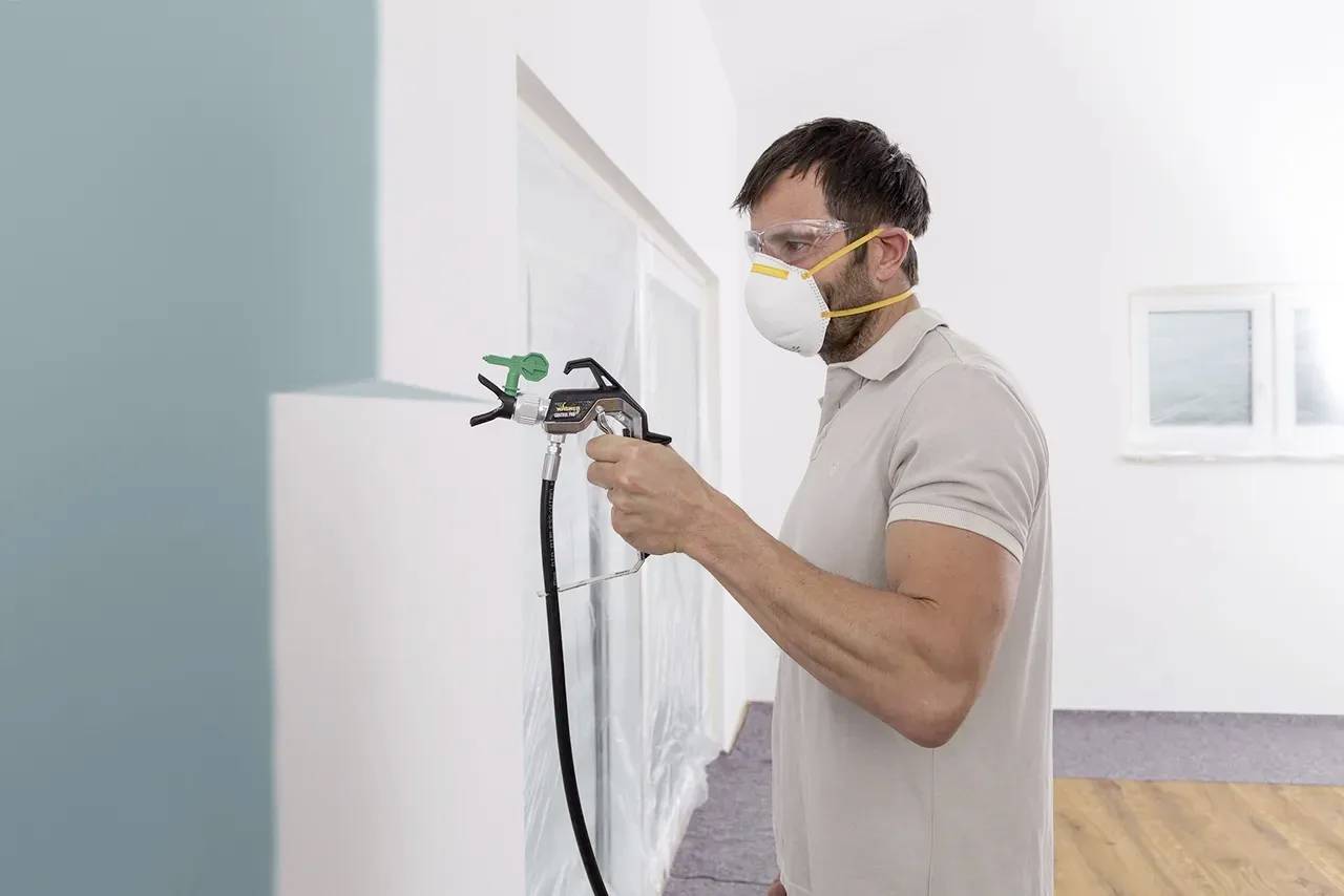 Ein Mann trägt Maske und Schutzbrille, während er mit einer Sprühpistole eine Wand in einem Innenraum streicht.