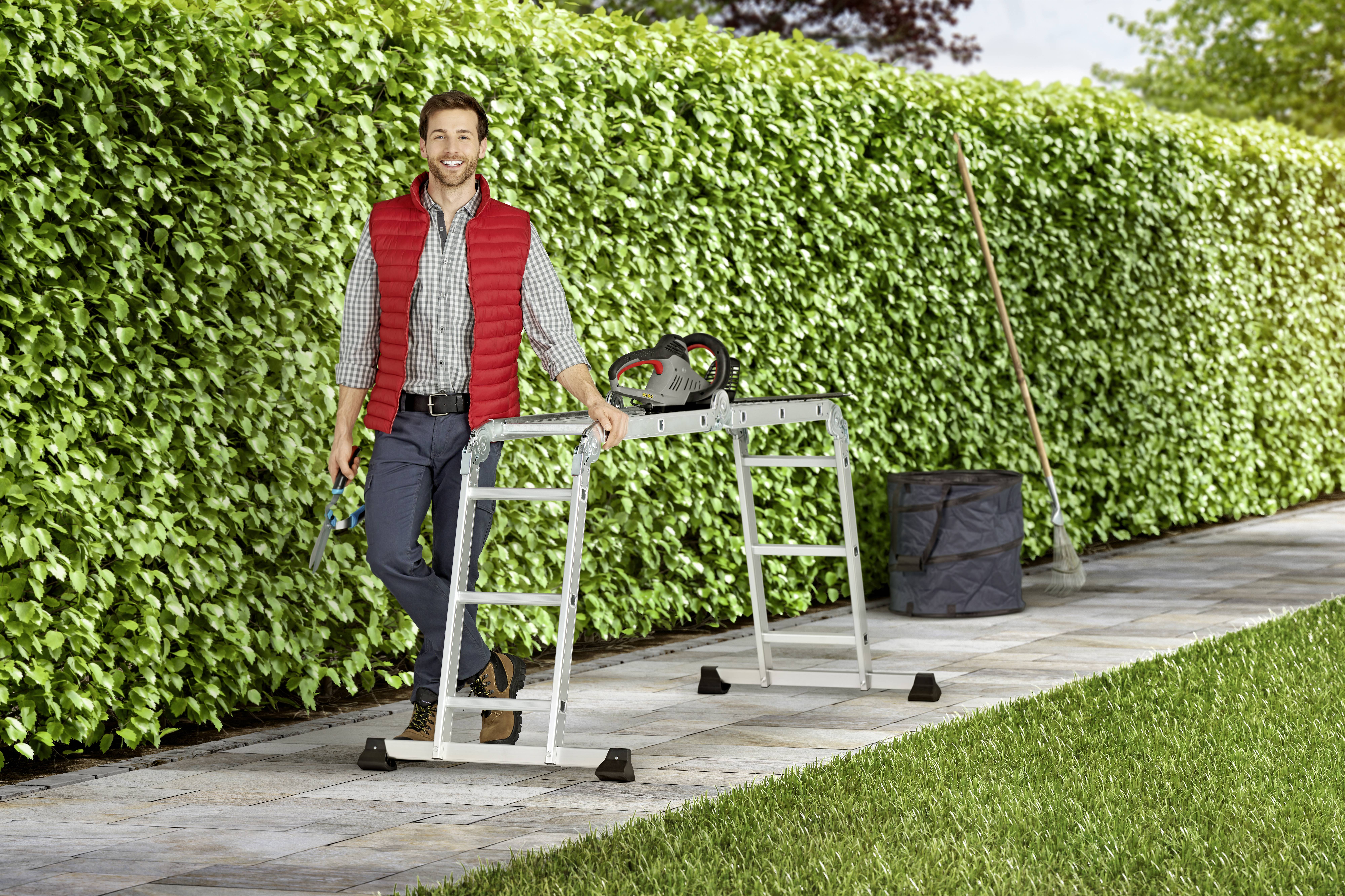 Eine Person lehnt an einer Metallleiter vor einer dichten, grünen Hecke und hält Gartenwerkzeuge. Ein Rechen und eine Tasche sind in der Nähe.