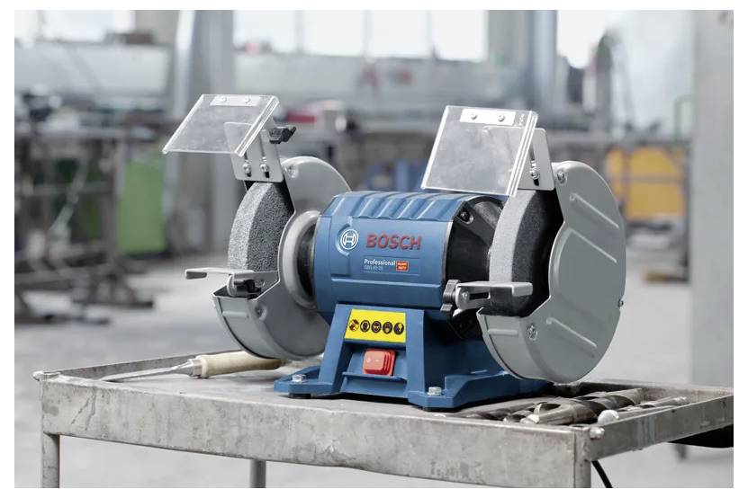 Eine Bankschleifmaschine mit einem 'Bosch'-Etikett steht auf einer Werkbank in einer Werkstatt. Die Maschine verfügt über Schutzschilde über jedem Schleifrad.