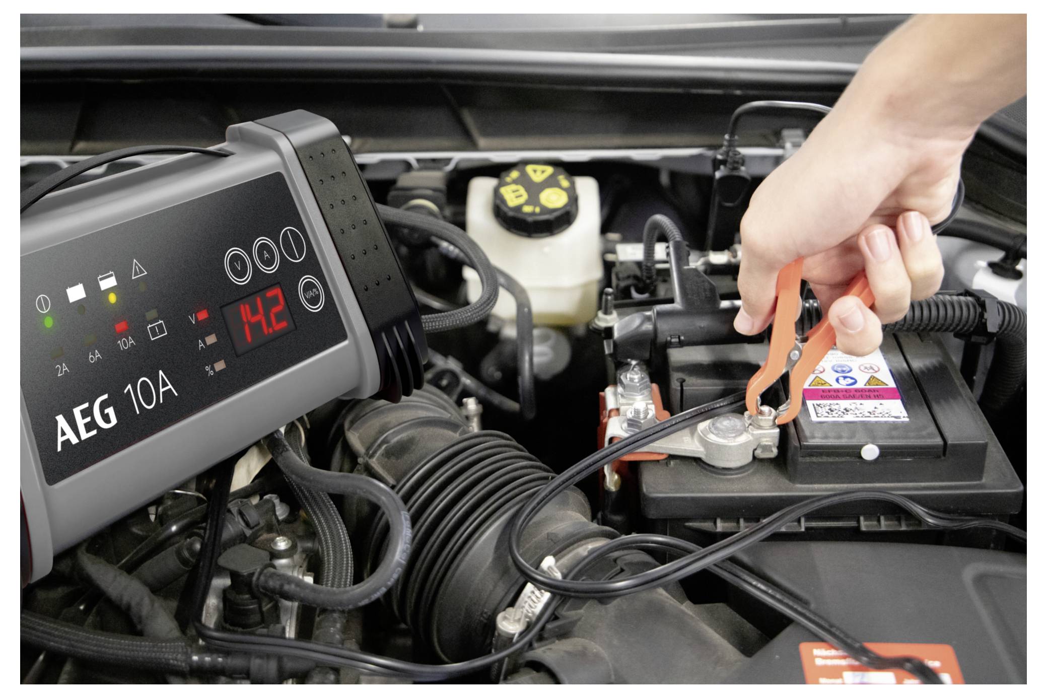 Die Hand einer Person befestigt eine Klemme eines Batterieladegeräts an einem Autobatterie-Anschluss. Das Ladegerät zeigt „14,2" an und trägt die Aufschrift „AEG 10A".