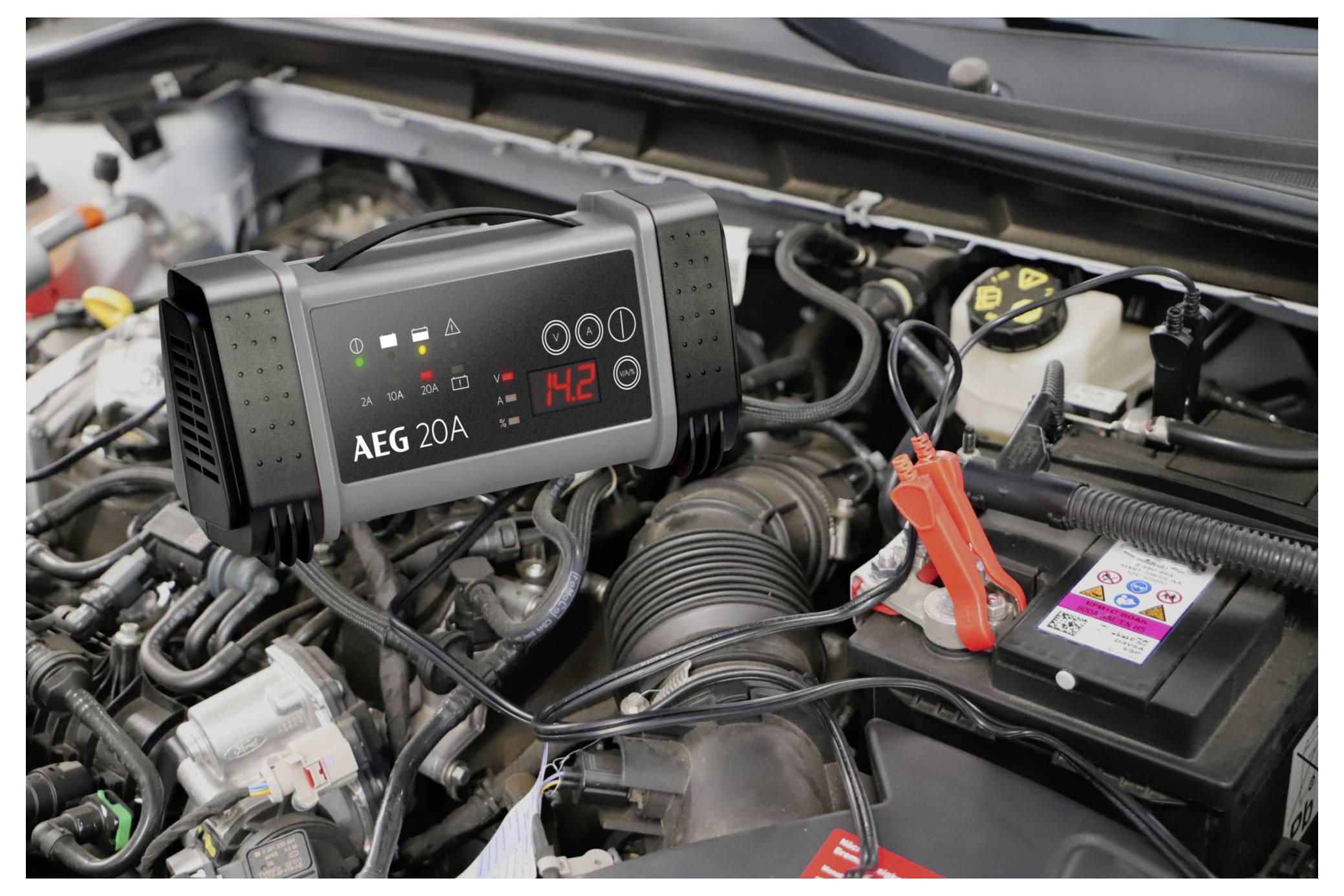 Ein Autobatterieladegerät, das "14,2" anzeigt, ist mit einer Autobatterie im Motorraum eines Fahrzeugs verbunden, wobei Kabel an den Batterieanschlüssen befestigt sind.