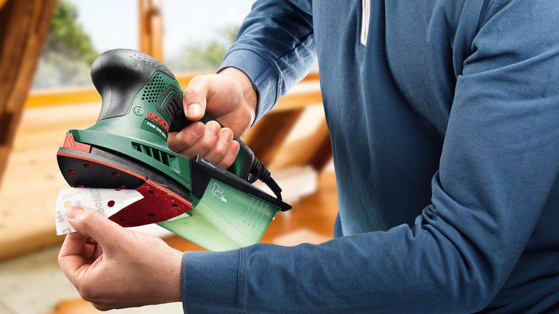 Eine Person nutzt einen grünen Elektroschleifer mit roter Schleiffläche, hält ein Stück Schleifpapier. Hintergrund zeigt Holzarbeiten.
