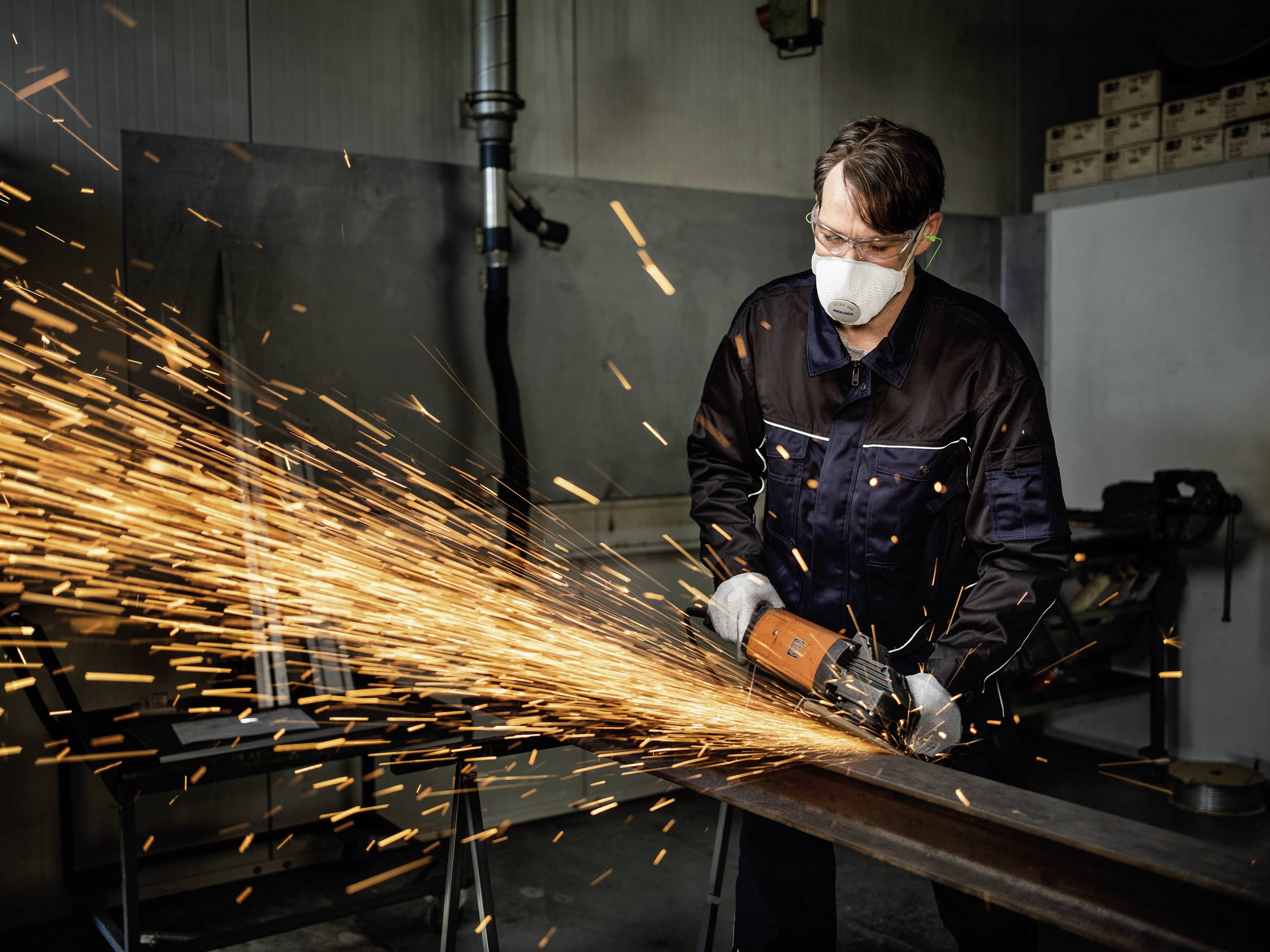 Ein Handwerker mit Schutzbrille und Maske schleift Metall in einer Werkstatt, wobei Funken sprühen.