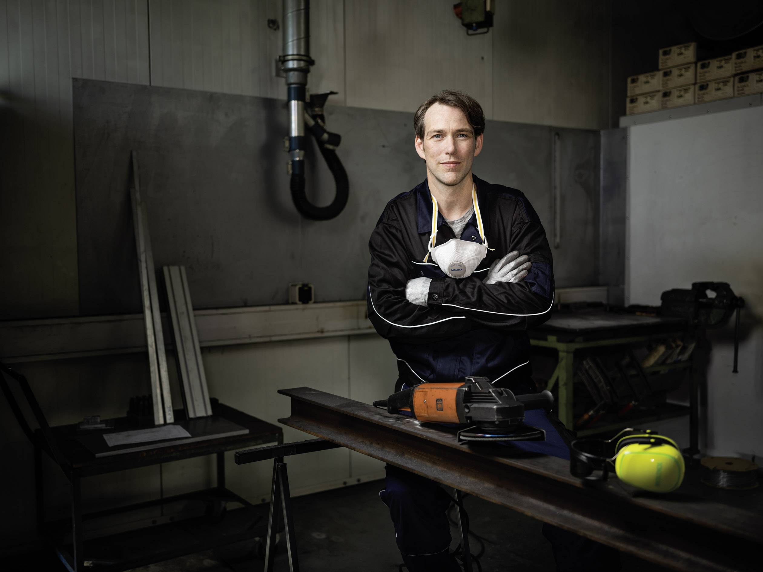 Ein Mann in Arbeitskleidung steht in einer Werkstatt neben einem Tisch mit einer Schleifmaschine und Gehörschutz.