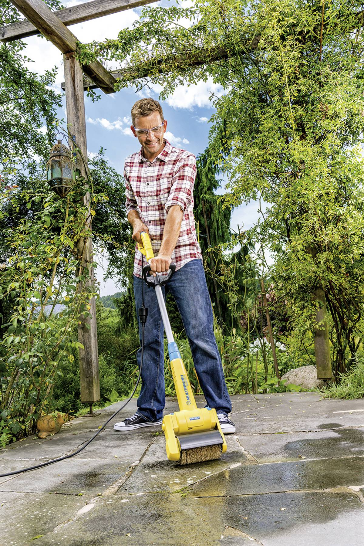 Eine Person reinigt eine Terrasse mit einem elektrischen Reinigungsgerät. Umgeben von üppigem Grün, unter einem Holzrahmen im Freien.