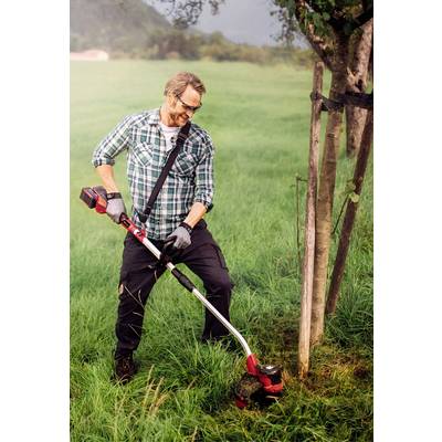 Ein Mann in kariertem Hemd und Sonnenbrille trimmt mit einem elektrischen Rasentrimmer Gras um einen Baum auf einer Wiese.