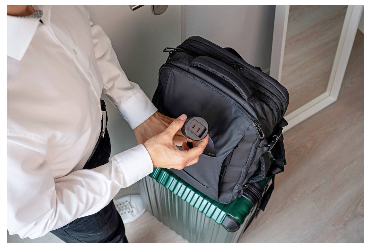 Eine Person hält ein rundes Verfolgungsgerät zur vorderen Tasche eines grünen Koffers neben einer schwarzen Reisetasche, was auf Gepäckverfolgung oder Identifizierung hindeutet.
