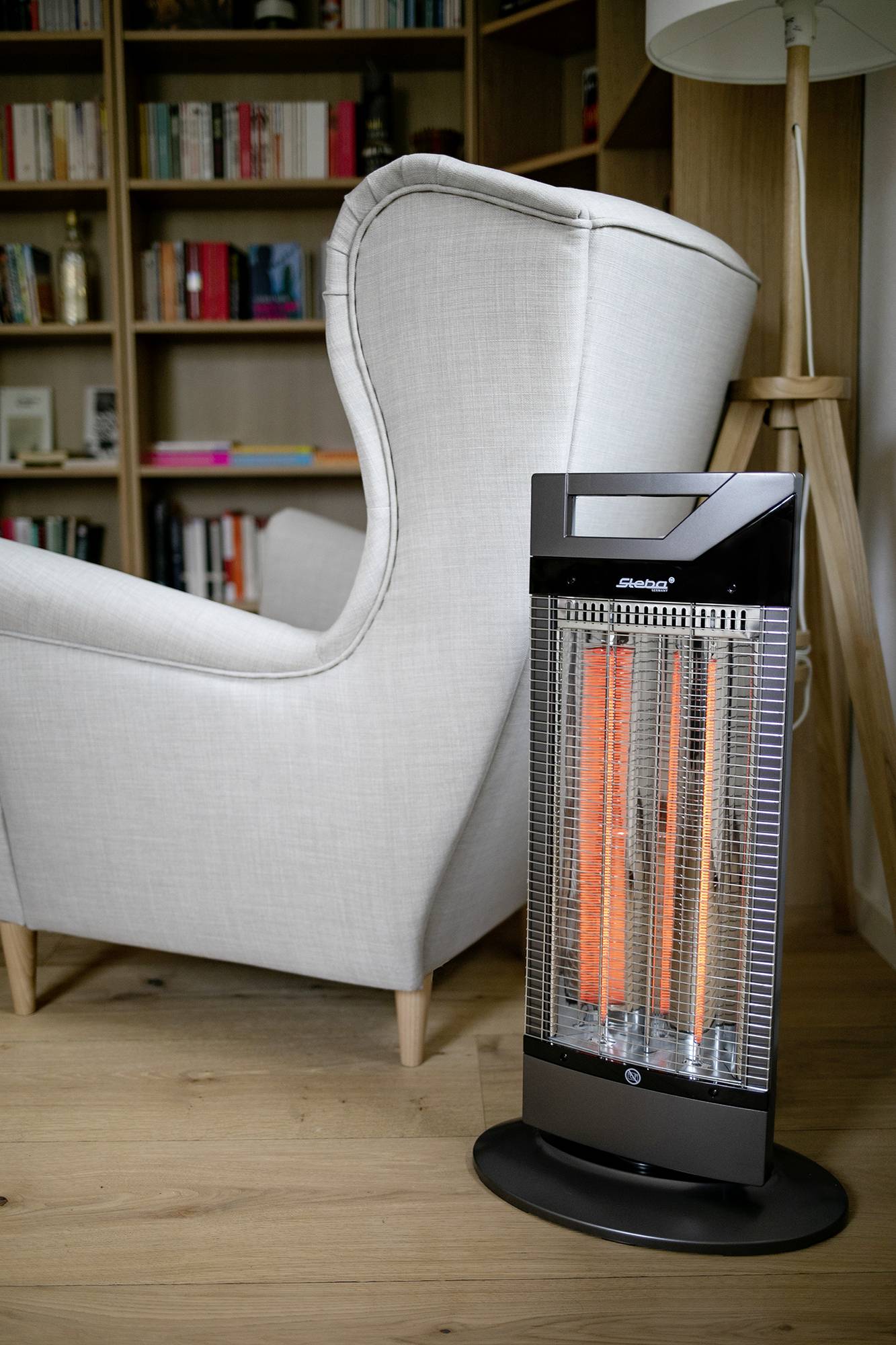 Ein grauer Sessel steht neben einem eingeschalteten Heizstrahler in einem Wohnzimmer mit Bücherregal im Hintergrund.