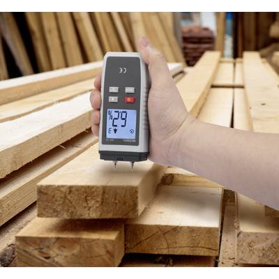 Eine Hand hält ein Holzfeuchtigkeitsmesser, der 29% anzeigt, über einem Stapel Holzlatten in einer Werkstatt.