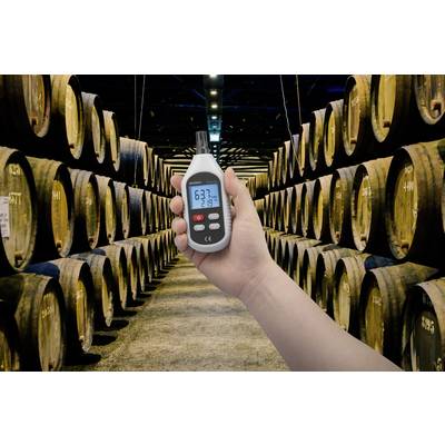 Eine Hand hält ein Hygrometer, das 63,7 % Luftfeuchtigkeit und 21,8 °C anzeigt. Im Hintergrund sind Holzfässer in einem Lagerraum zu sehen.