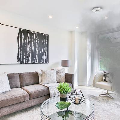 Ein moderner Wohnraum mit einem braunen Sofa, einem Glastisch, abstrakter Kunst an der Wand und viel natürlichem Licht durch ein großes Fenster.
