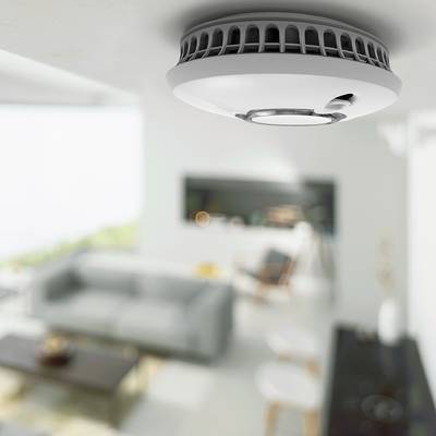 Ein Rauchmelder ist an der Decke eines modernen Wohnzimmers montiert. Im Hintergrund sind ein Sofa und ein Tisch zu sehen.