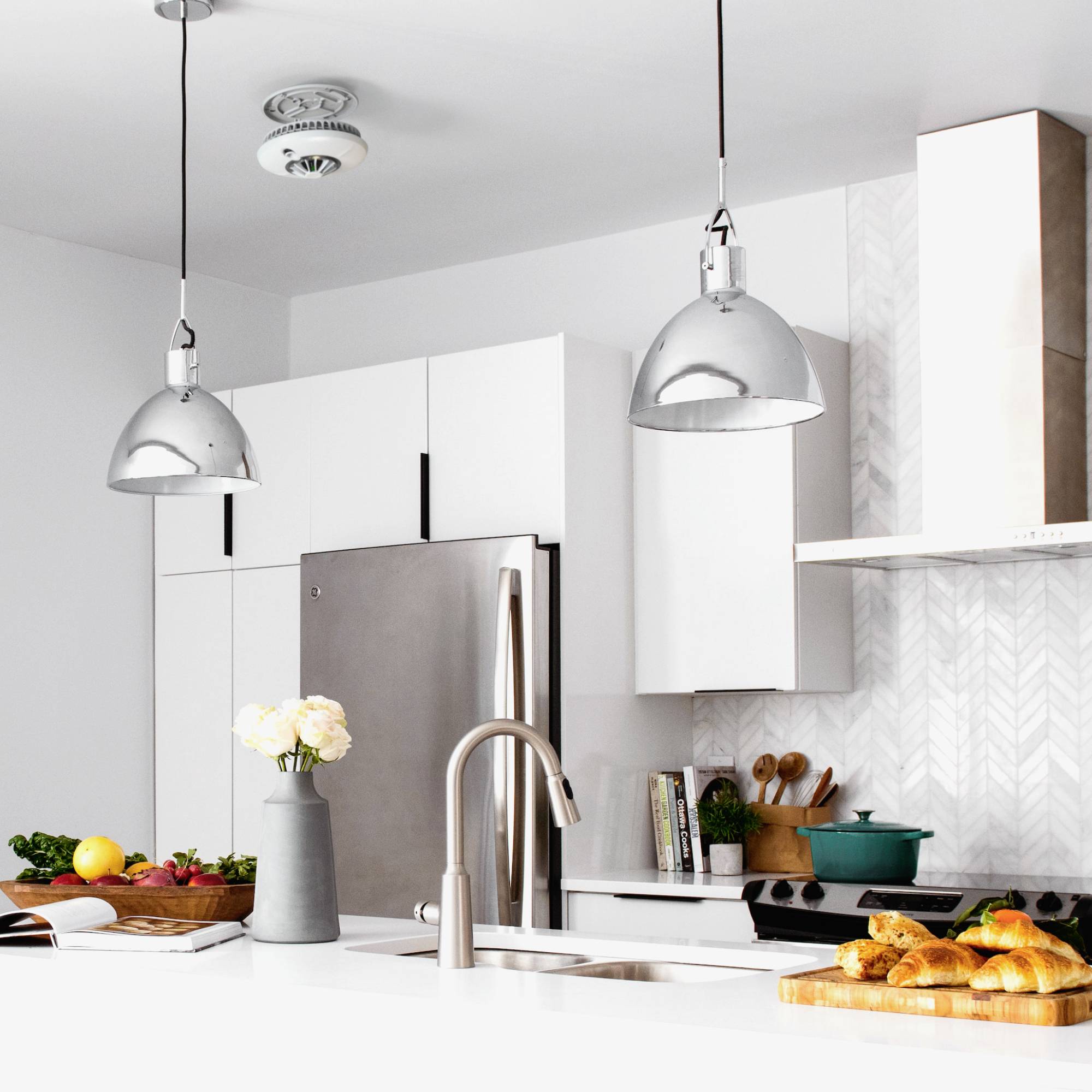 Helle, moderne Küche mit weißen Schränken, Metallleuchten, Obst auf dem Tisch und einem Edelstahlkühlschrank.
