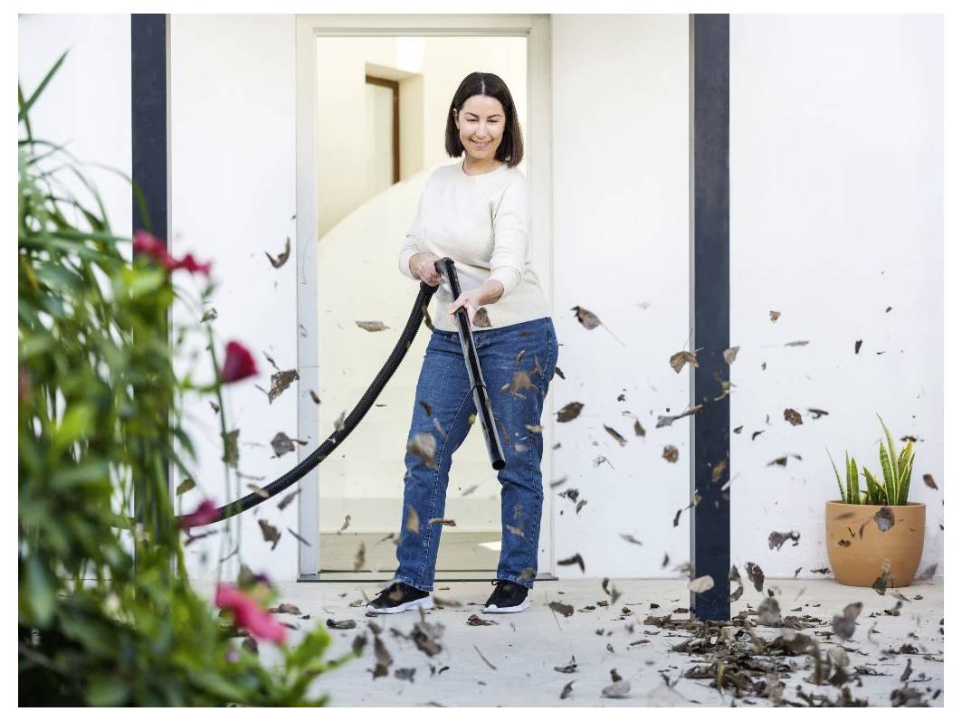 Eine Frau benutzt einen Laubbläser, um Blätter von einer Terrassenfläche zu entfernen. Sie steht in einer offenen Türöffnung eines weißen Gebäudes und lächelt.