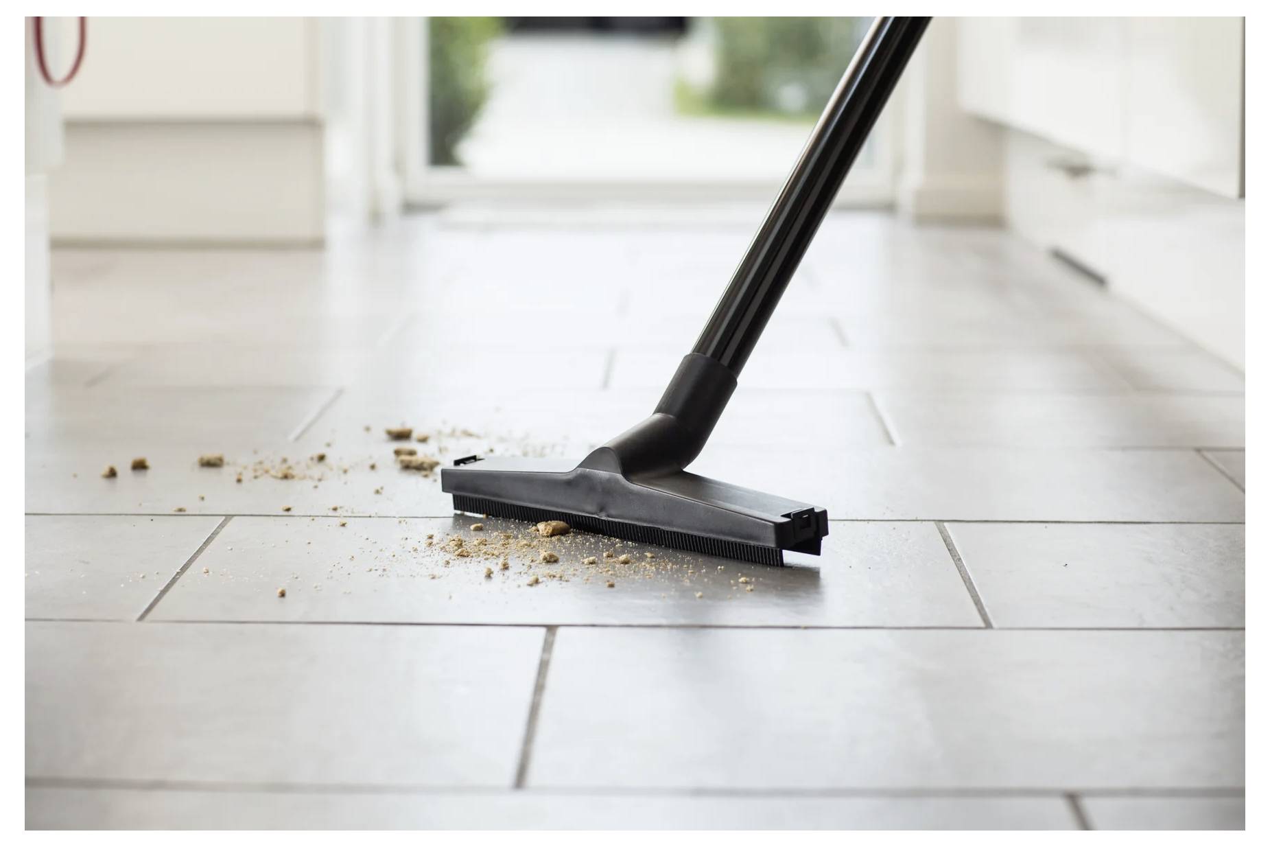 Ein Staubsauger entfernt Krümel von einem gefliesten Küchenboden und vermittelt damit einen Eindruck von Sauberkeit und Ordentlichkeit in einem häuslichen Umfeld.