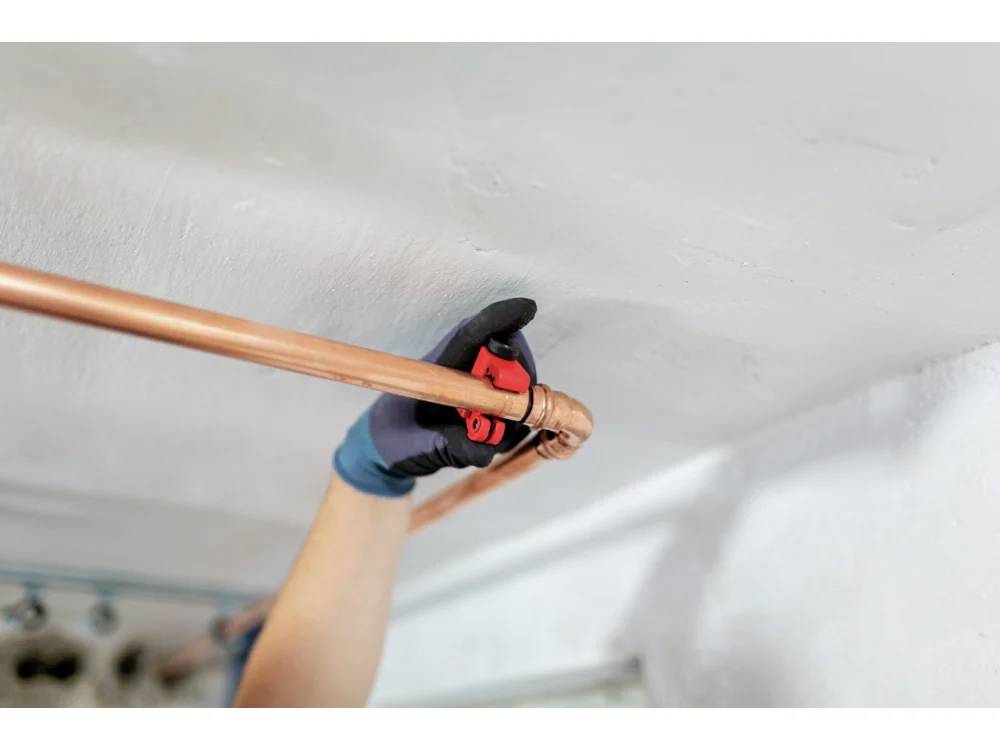 Eine Person mit Handschuhen zieht eine Kupferrohrleitung unter einer weißen Decke mit einem roten Werkzeug fest, was auf eine Klempner- oder Wartungsaufgabe hindeutet.