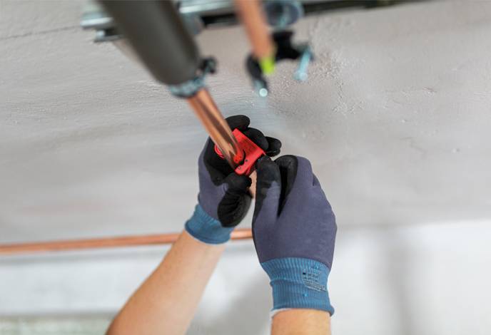 Eine Person mit Handschuhen repariert eine Kupferrohrleitung an der Decke und verwendet dabei einen Schraubenschlüssel oder ein ähnliches Werkzeug, wobei sie sich auf Sanitärreparaturen konzentriert.