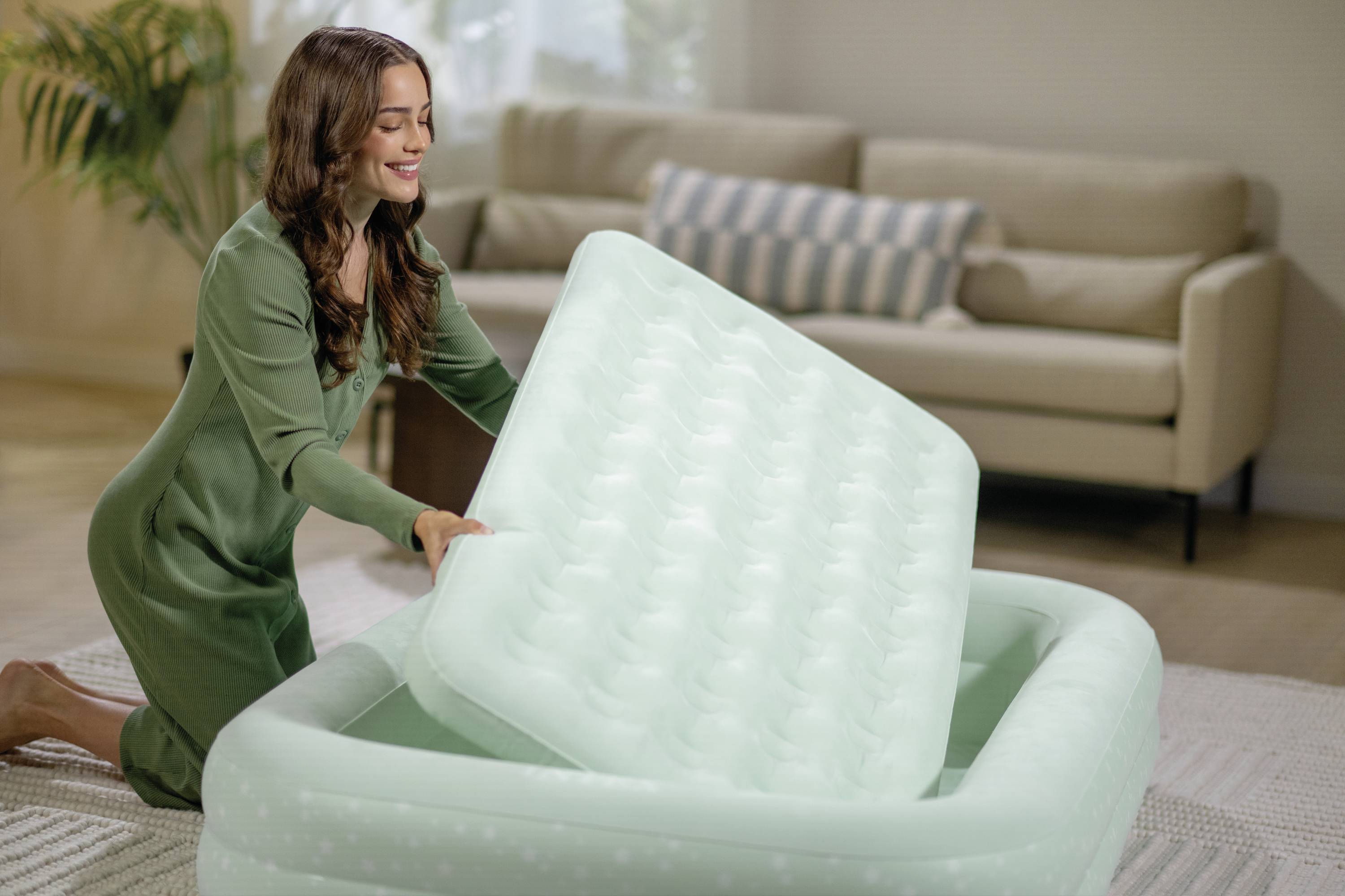 Eine Person in einem grünen Outfit richtet eine Luftmatratze in einem Wohnzimmer ein, in dem ein beiger Sofa und eine Pflanze im Hintergrund stehen.