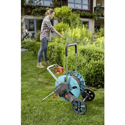 Eine Person gießt einen Garten mit einem Schlauch, der an einem blauen Schlauchwagen befestigt ist. Der Garten ist grün und blüht.