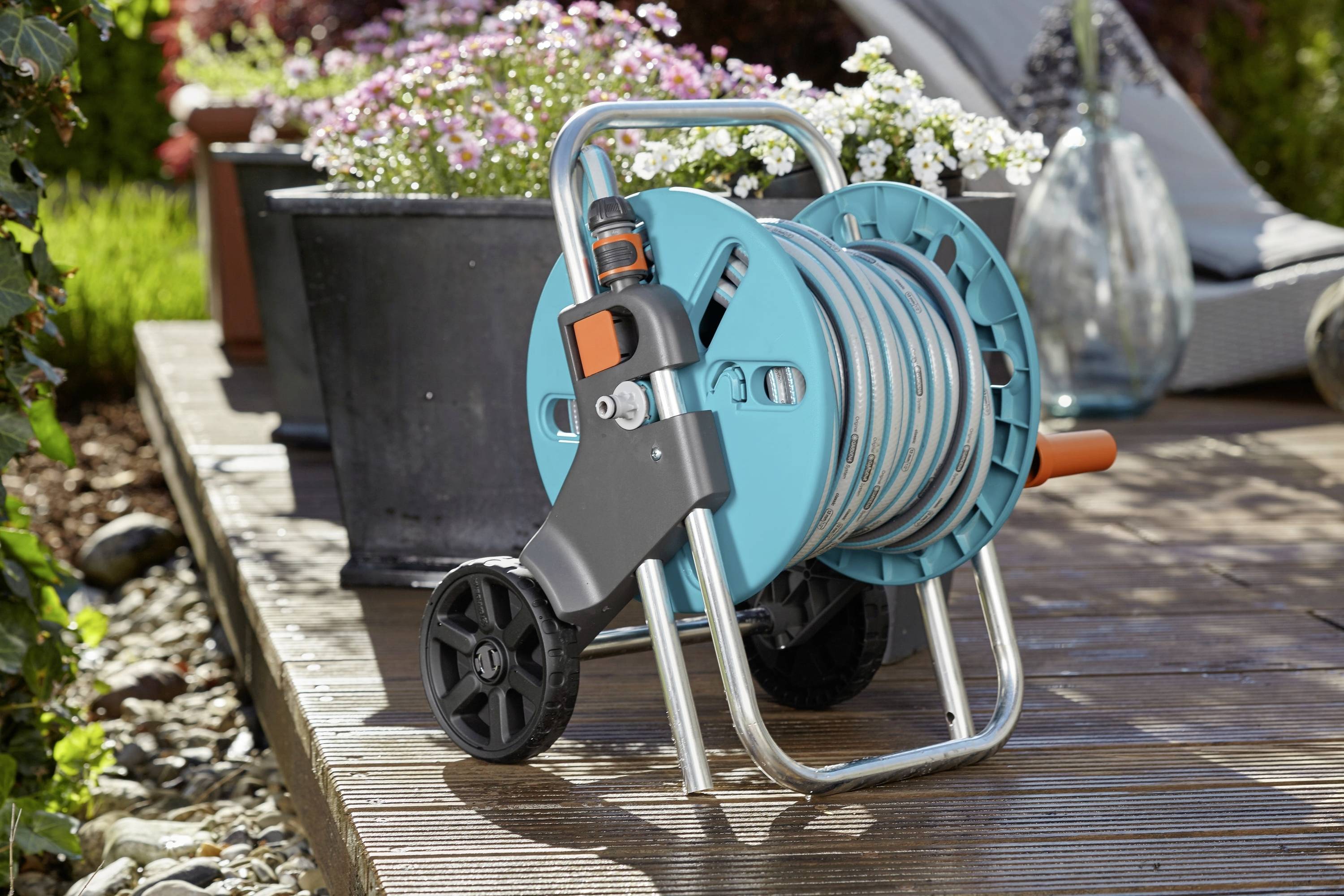 Ein Gartenschlauch auf einem Rollwagen befindet sich auf einer Terrasse neben blühenden Blumen in Töpfen.