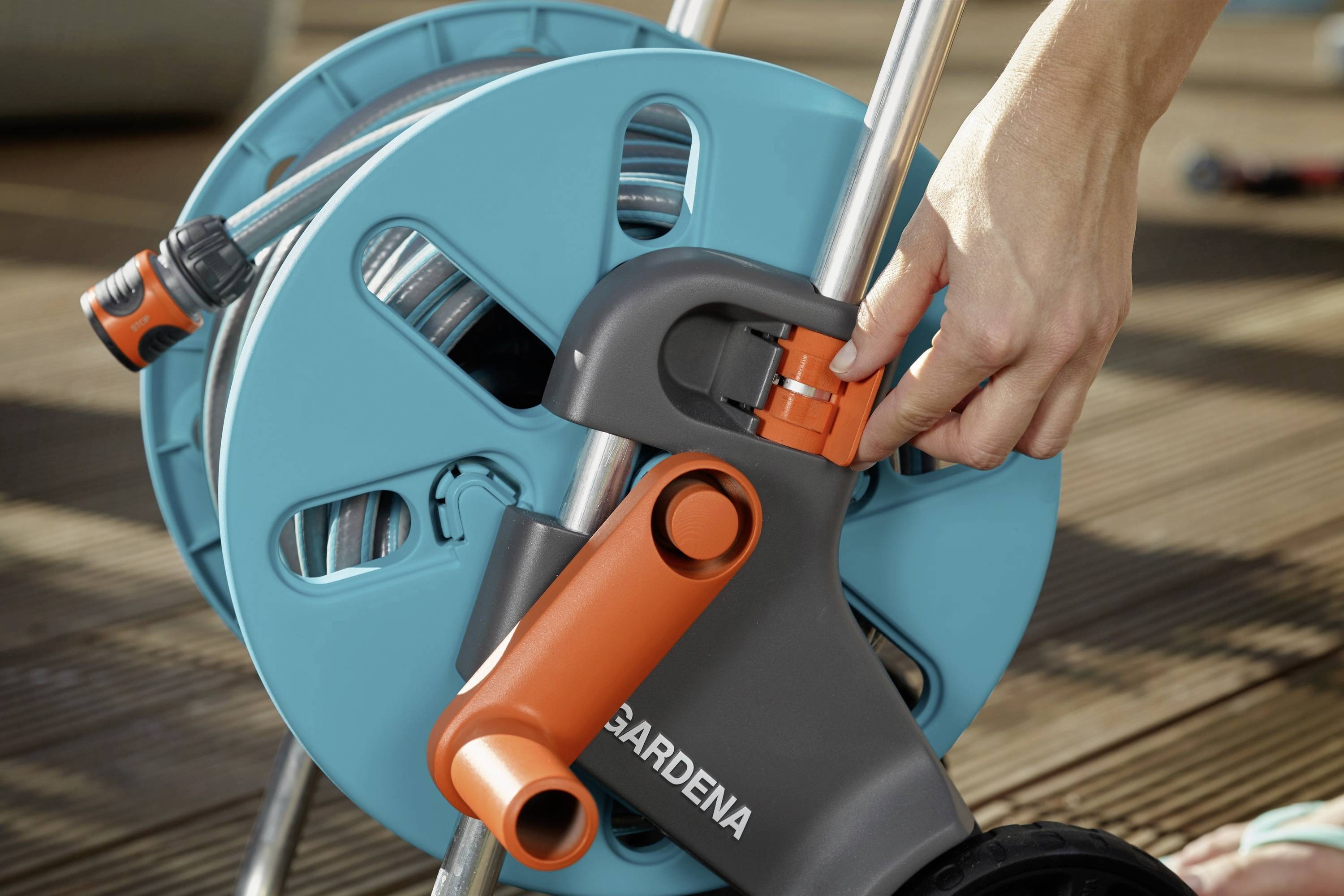 Eine Hand justiert den Schlauchanschluss an einem Gartenschlauchwagen. Der Schlauchwagen ist blau und orange und steht auf einer Holzterrasse.