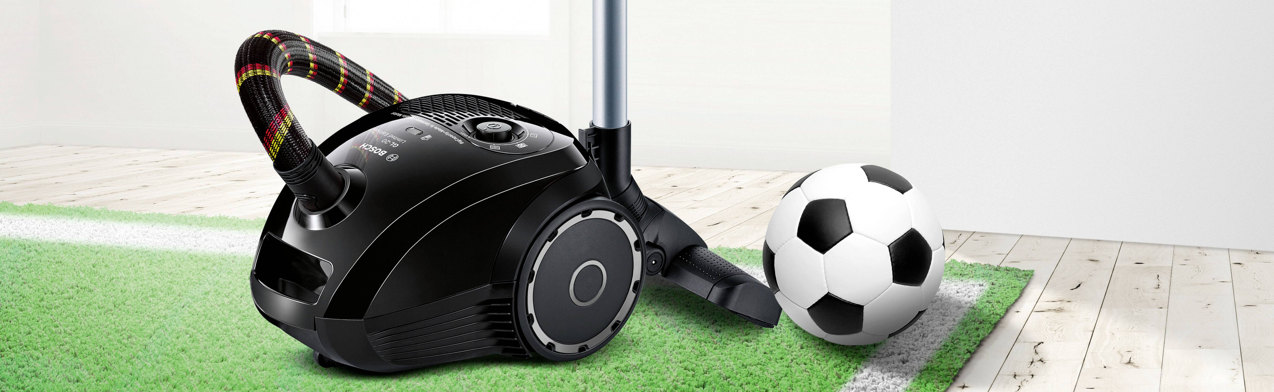 Ein schwarzer Staubsauger steht auf einem grünen Teppich neben einem Fußball. Im Hintergrund ist ein Holzboden und eine weiße Wand zu sehen.