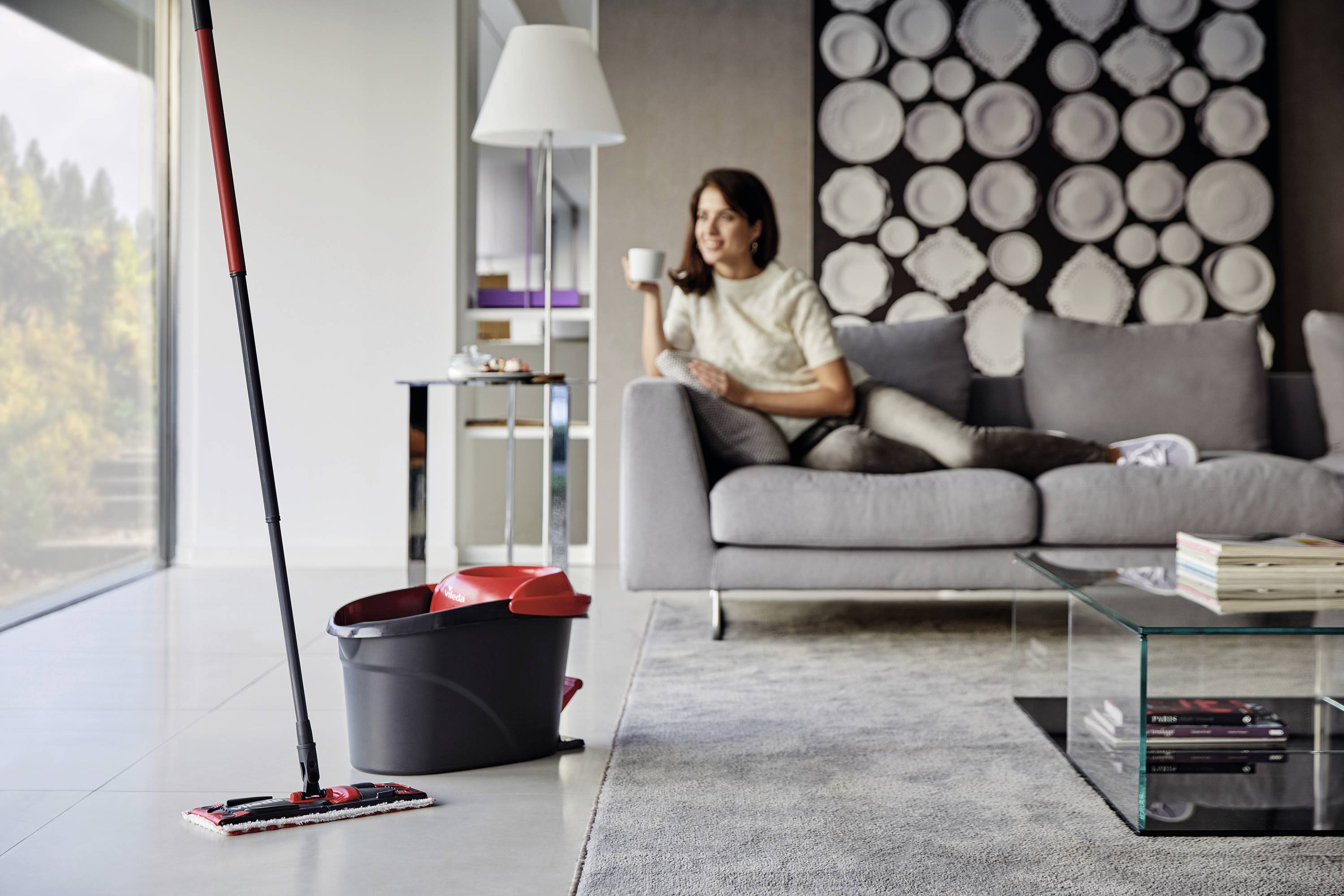 Frau sitzt entspannt auf einem Sofa in einem modern eingerichteten Wohnzimmer mit einem Wischmop und Eimer im Vordergrund.