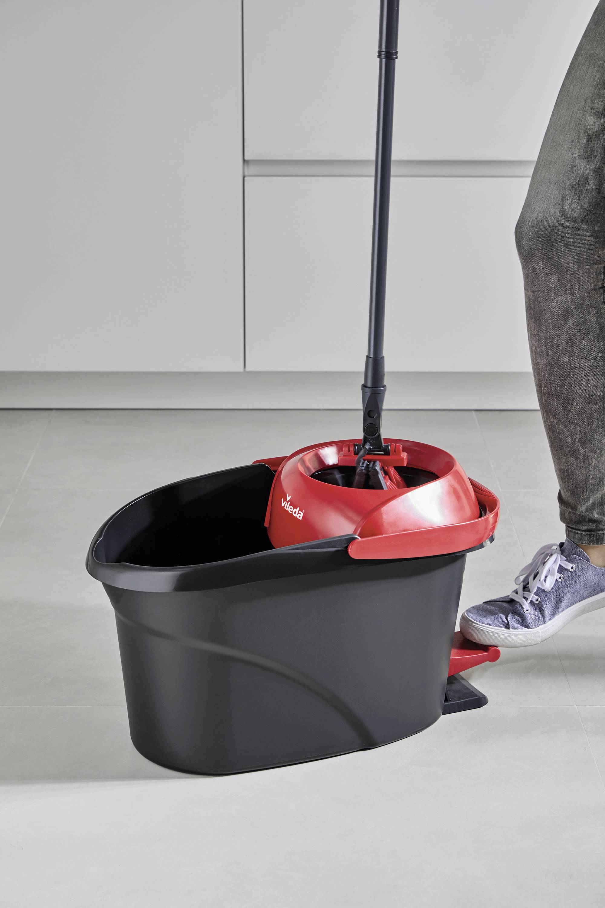 Eine Person wischt den Boden mit einem schwarzen Eimer und einem roten Wringaufsatz, während sie helle Turnschuhe trägt.